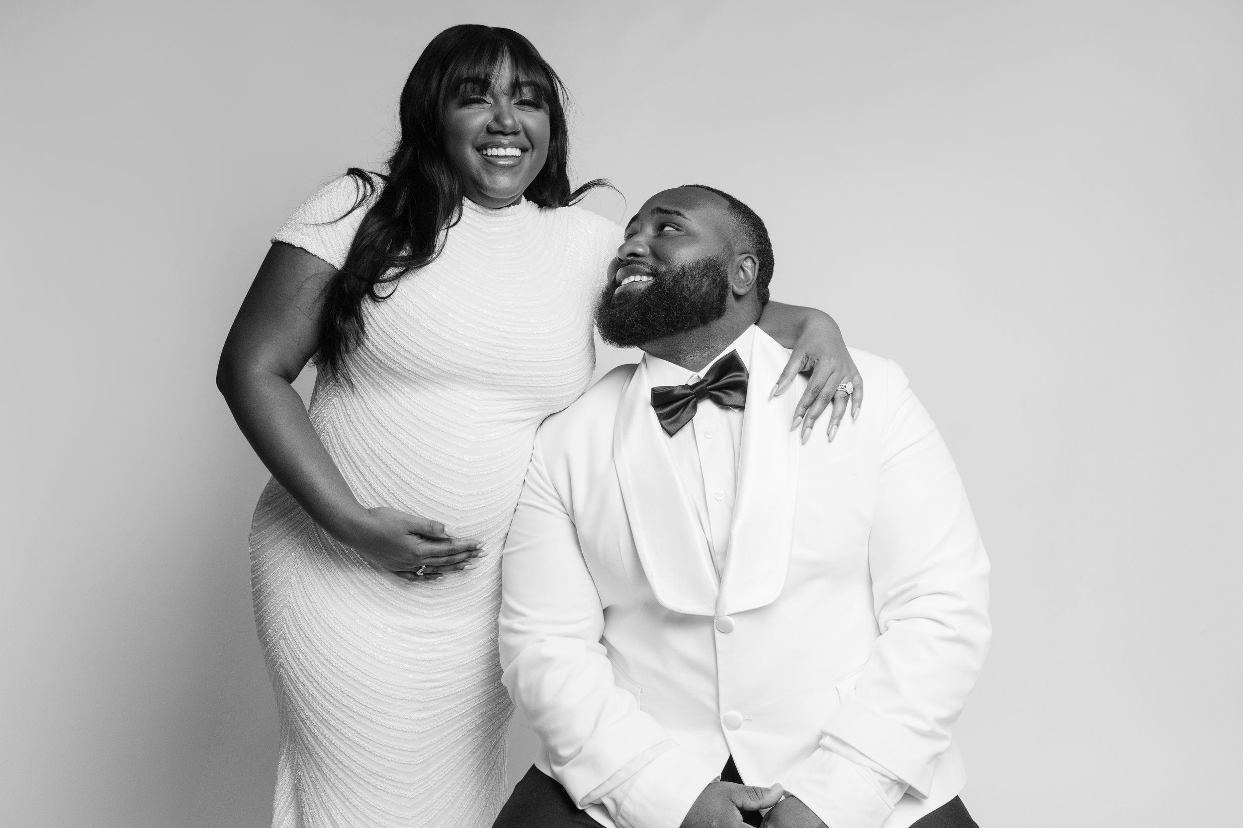 Black and white photo of a smiling couple in white formal wear, woman in gown standing beside seated bearded man in tuxedo.
