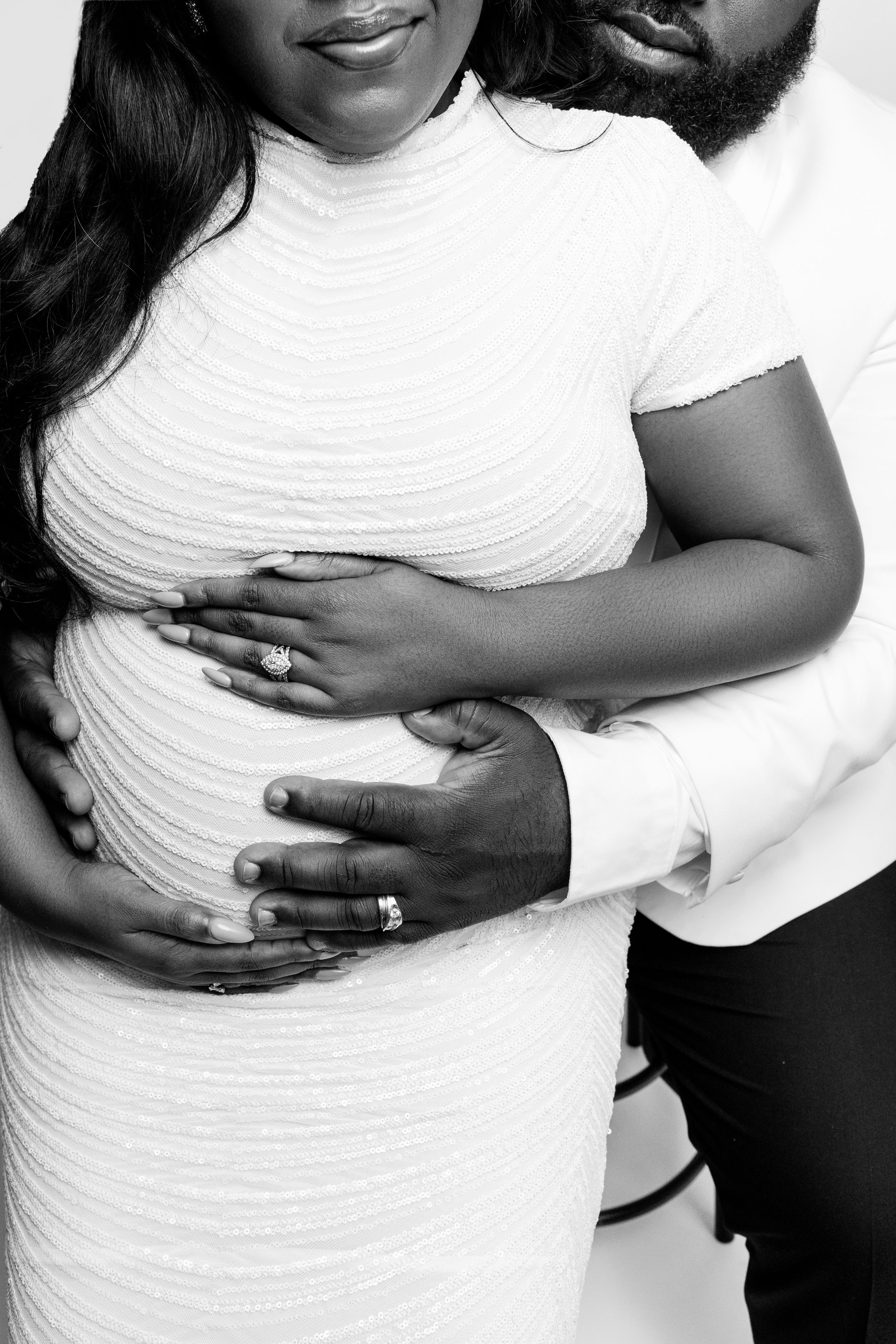 Black and white maternity photo of a couple in white outfits, hands cradling pregnant belly.