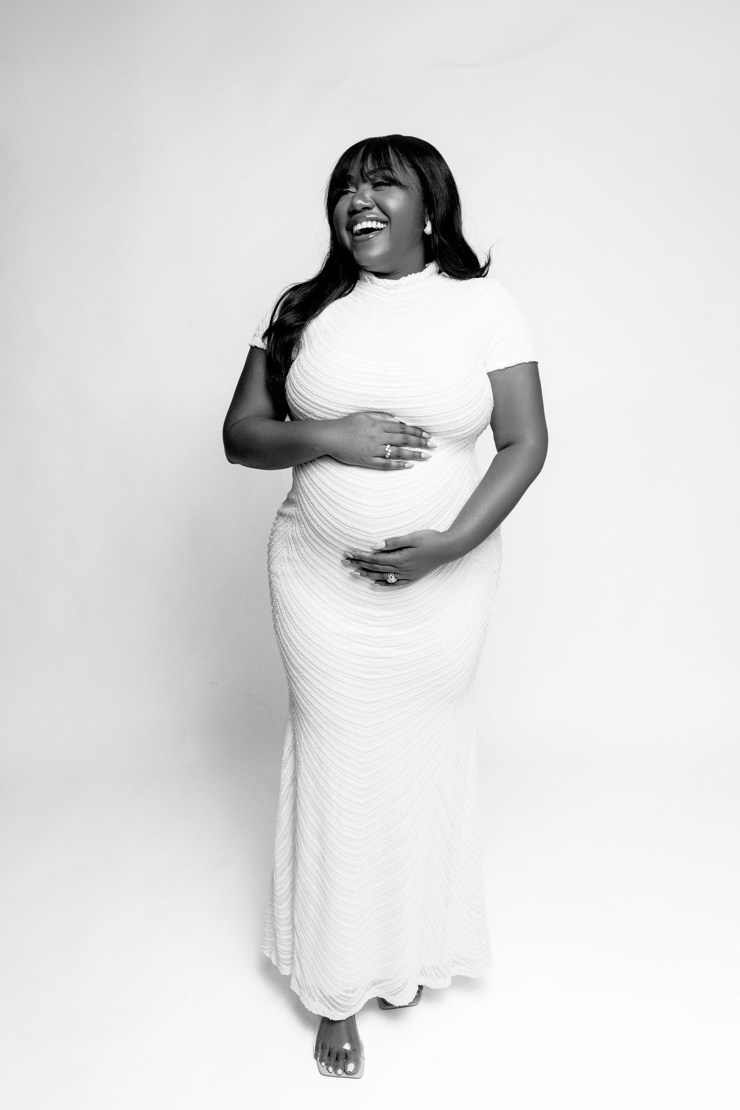 Smiling pregnant woman in white ribbed maxi dress cradling baby bump, black and white maternity portrait photo.