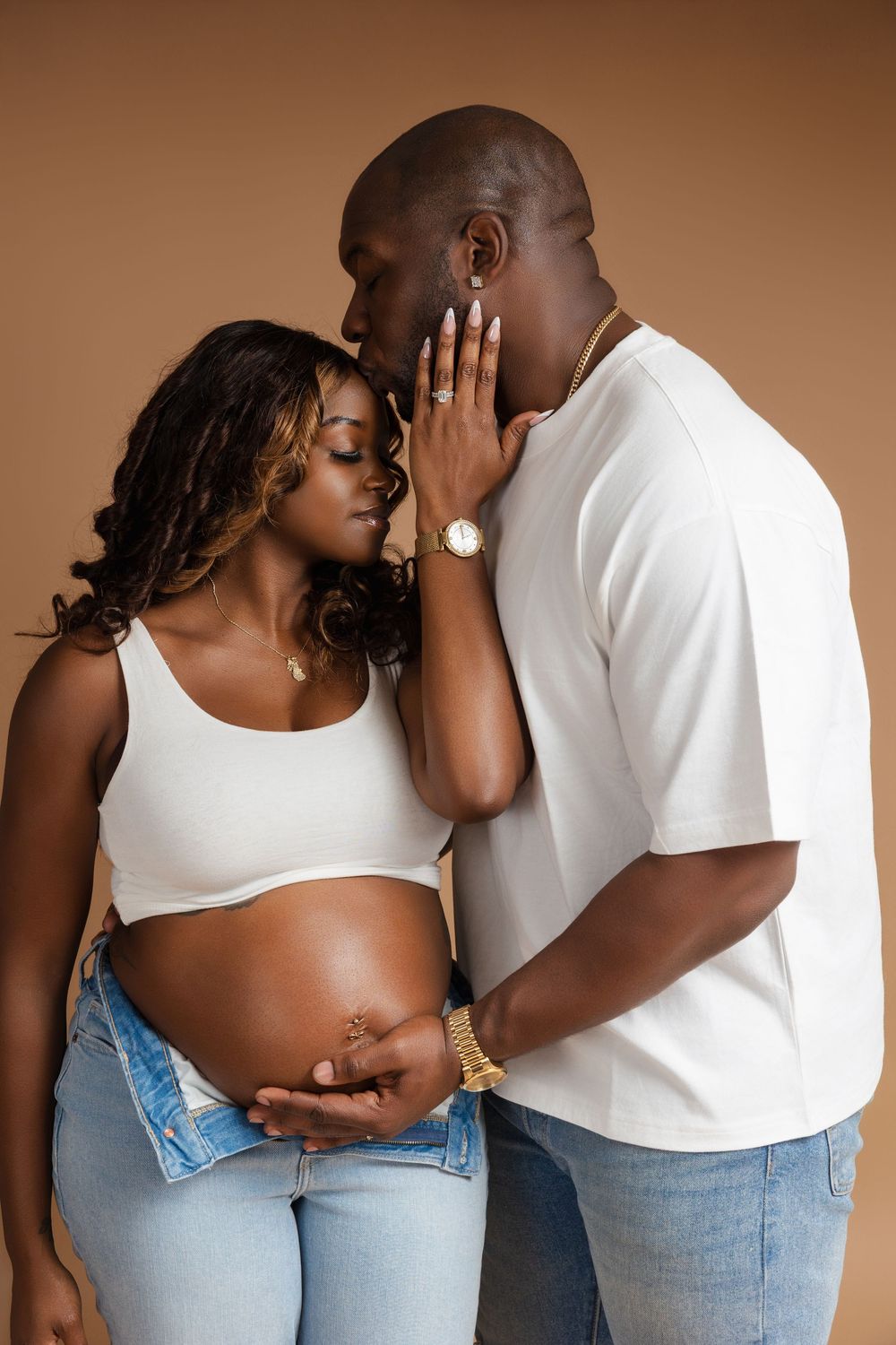 Expectant couple shares an intimate moment against brown backdrop, dressed in white tops and denim.