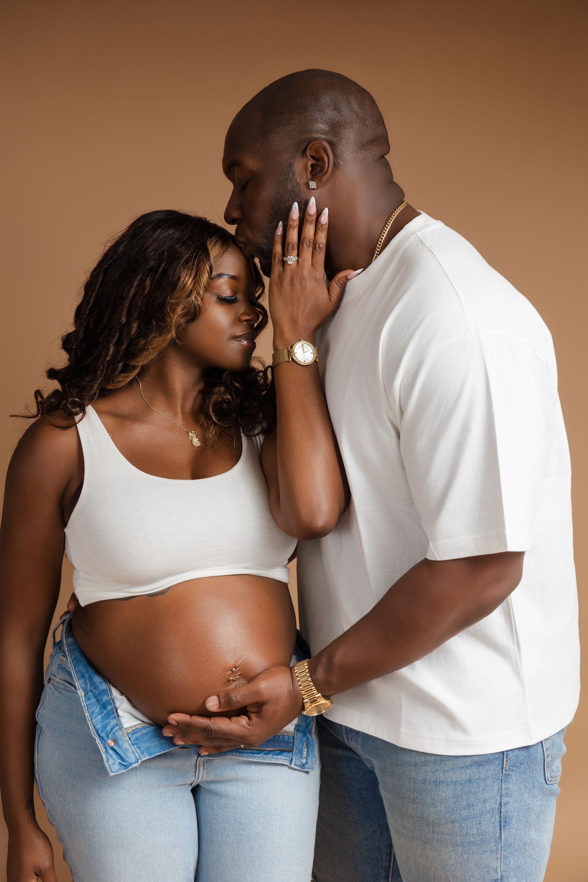 Expectant couple shares an intimate moment against brown backdrop, dressed in white tops and denim.