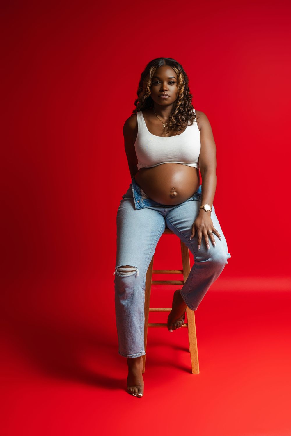 A pregnant woman in ripped jeans and white crop top poses confidently on a wooden stool against a vibrant red backdrop.