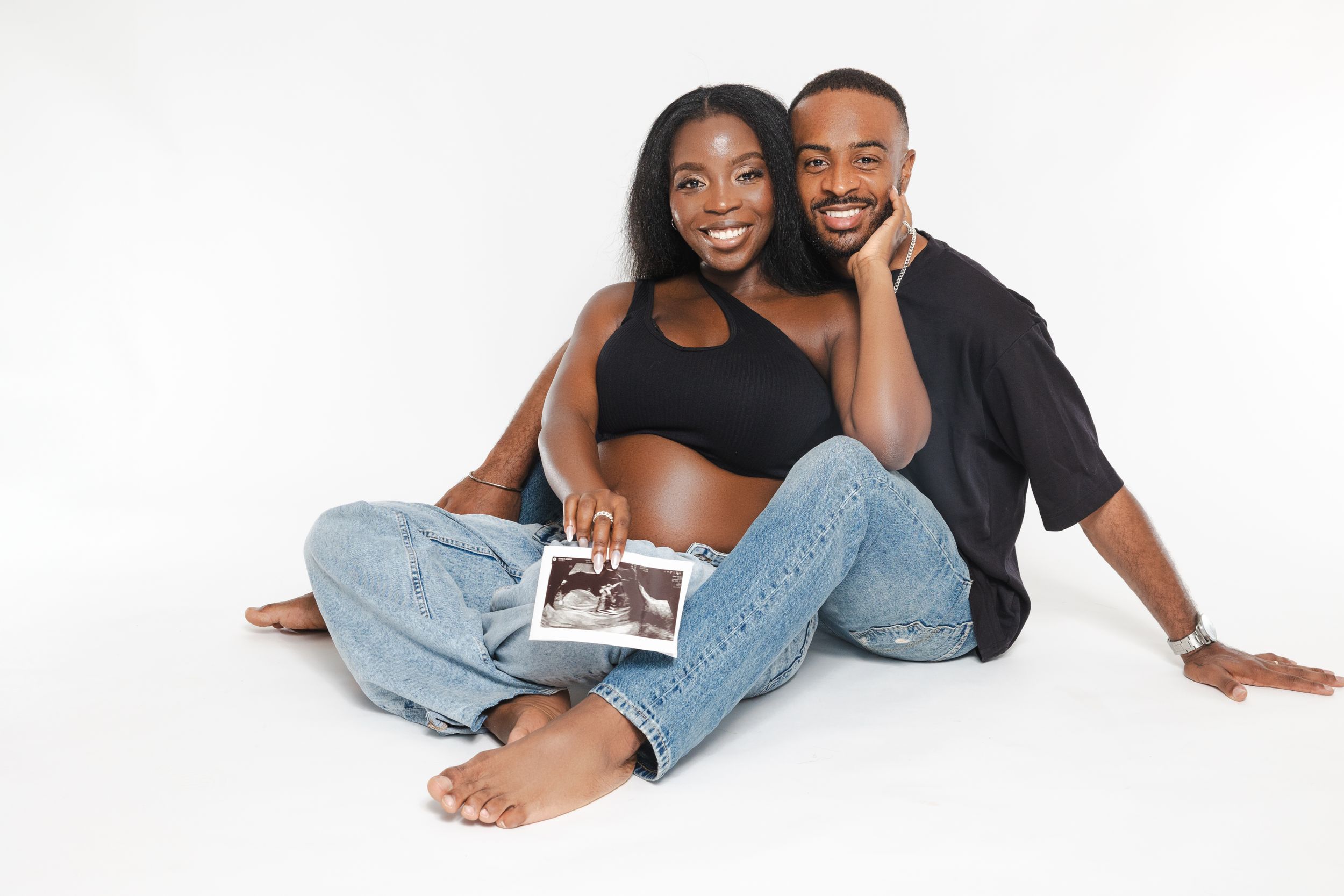 Couple sitting casually on white background holding ultrasound photo.