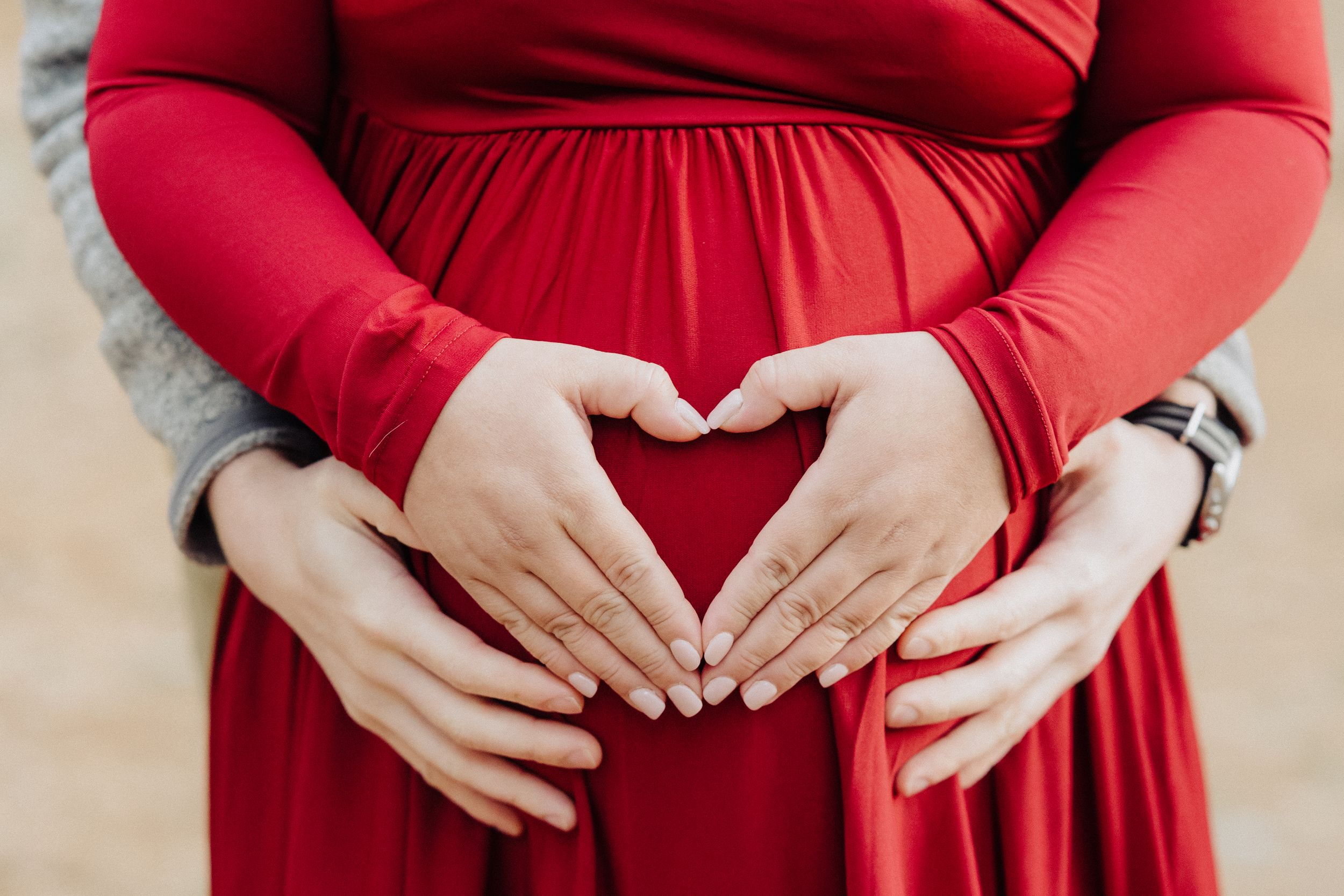 Close-up of hands forming heart shape over pregnant belly in red maternity dress.