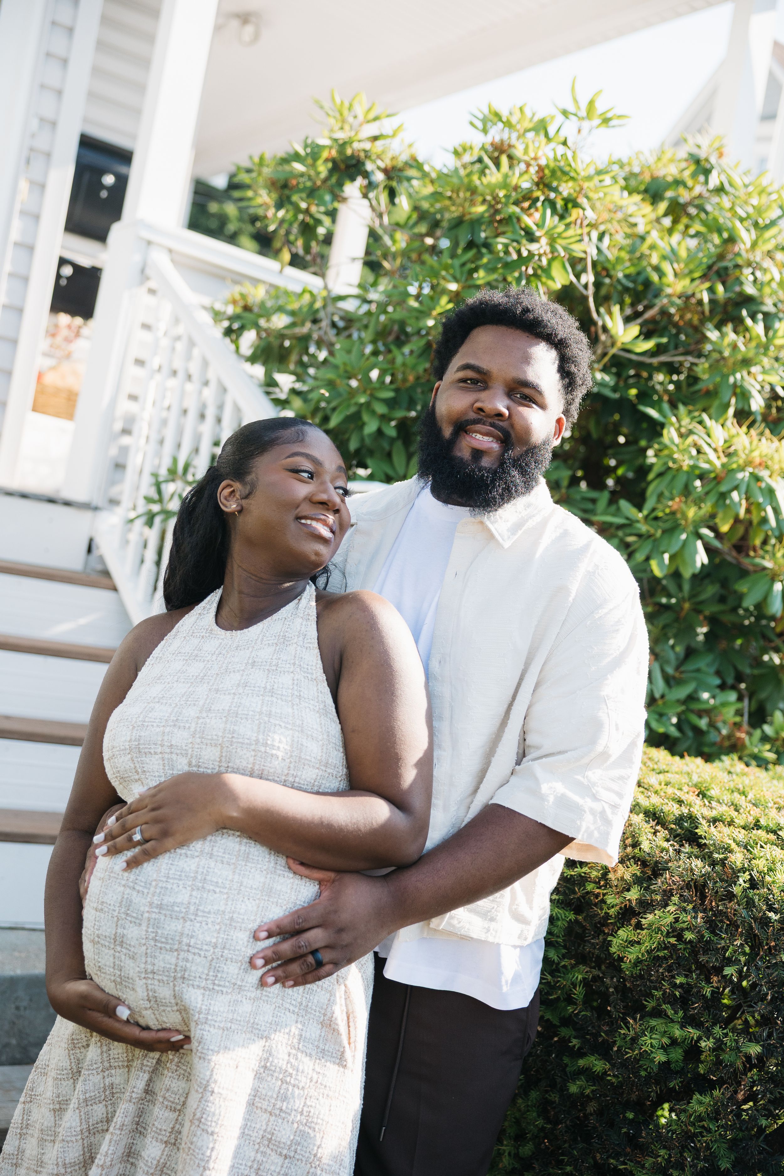 Happy pregnant couple embracing outdoors in front of a white porch and green bushes on a sunny day.