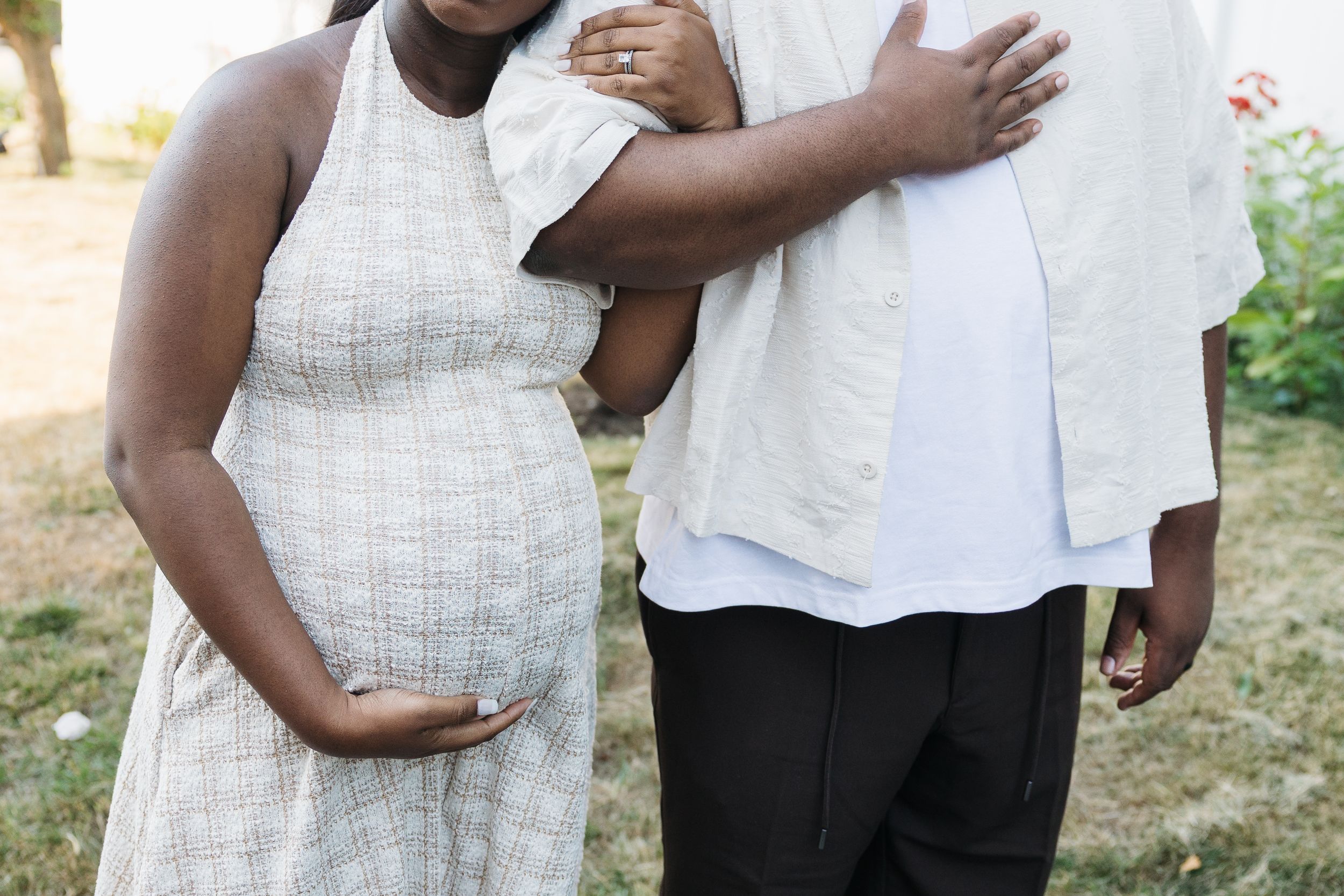 Pregnant Black woman in white dress standing with partner outdoors, both facing away, showing baby bump.