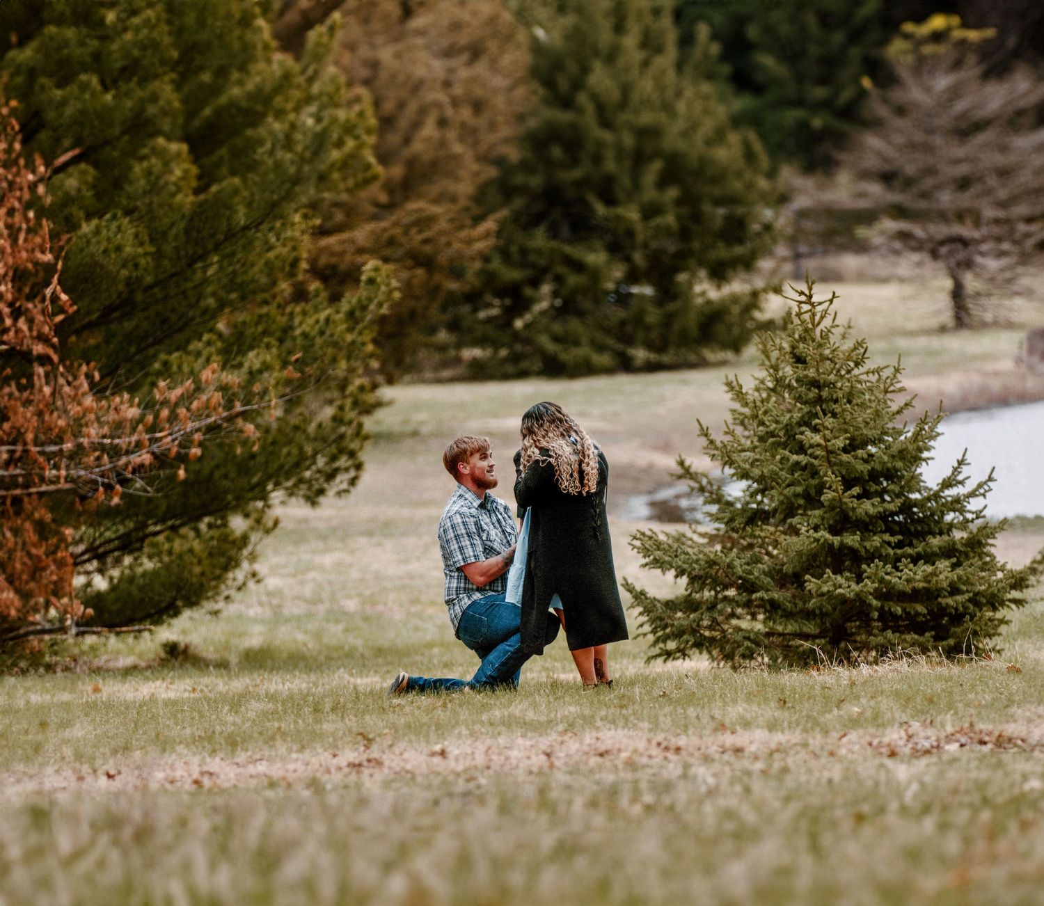 A romantic proposal moment between a couple near evergreen trees in an outdoor setting.