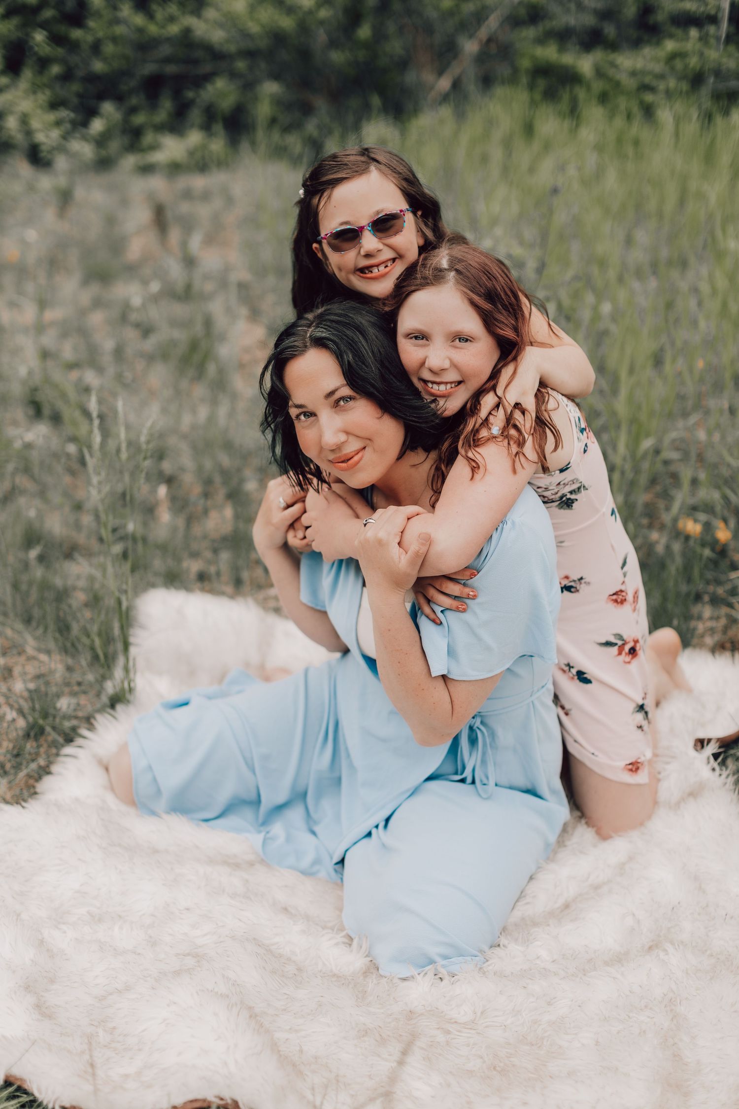 Three smiling girls hugging and posing together on a white blanket in a green meadow.
