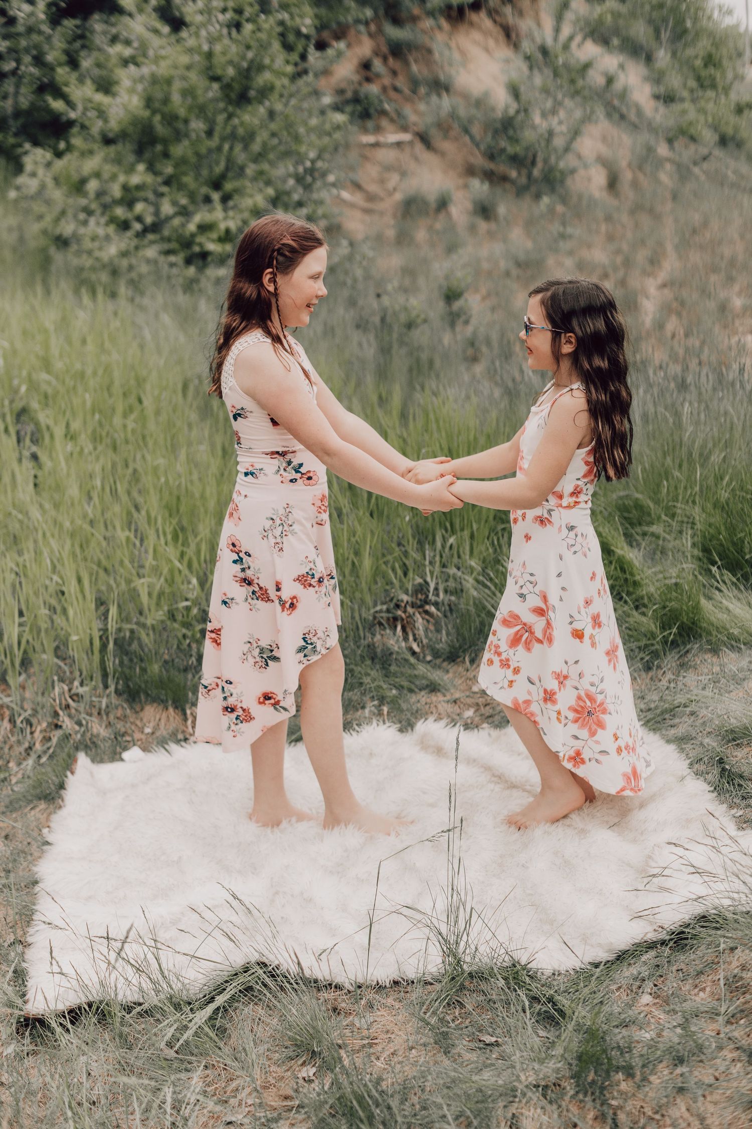 Two girls in floral dresses holding hands and spinning on a white blanket in a green meadow.