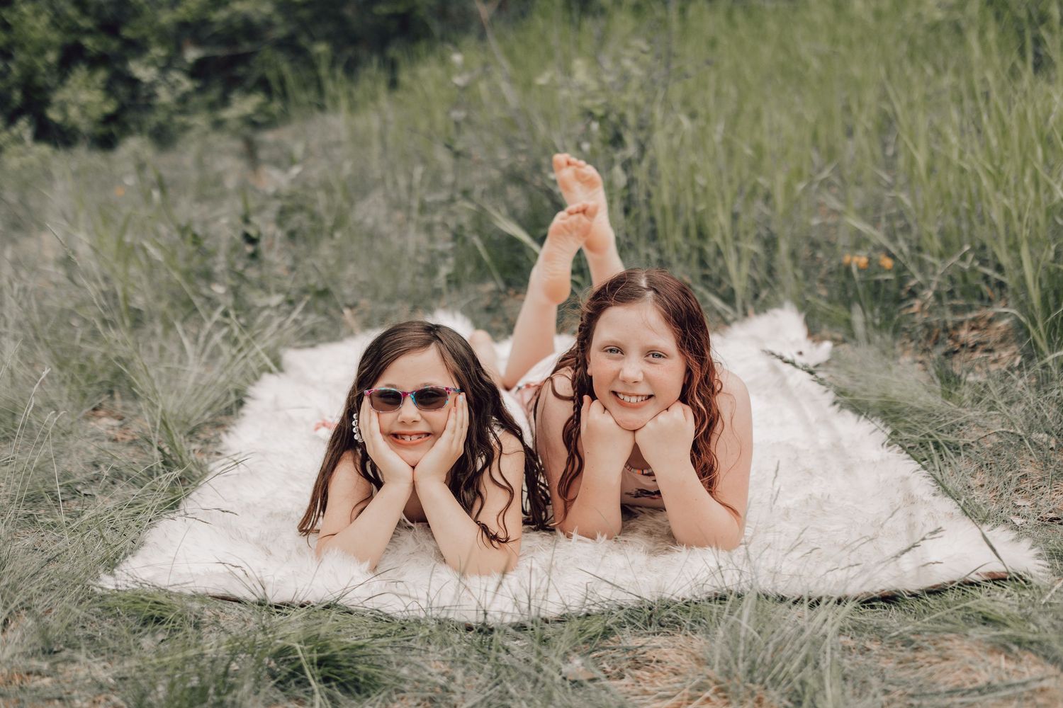 Two smiling girls lying on a white blanket in a grassy field, one wearing sunglasses.