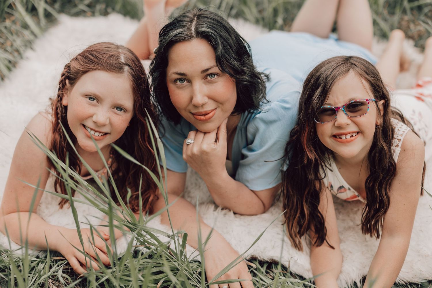 A mother and two daughters smiling while lying on a blanket surrounded by green grass.