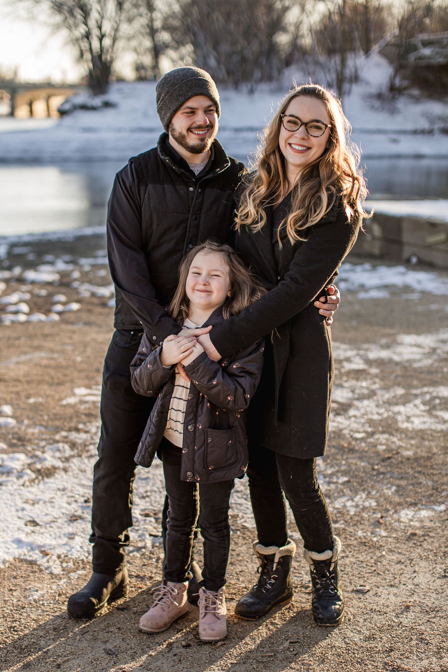 A young family of three posing together near a snowy riverbank on a sunny winter day.