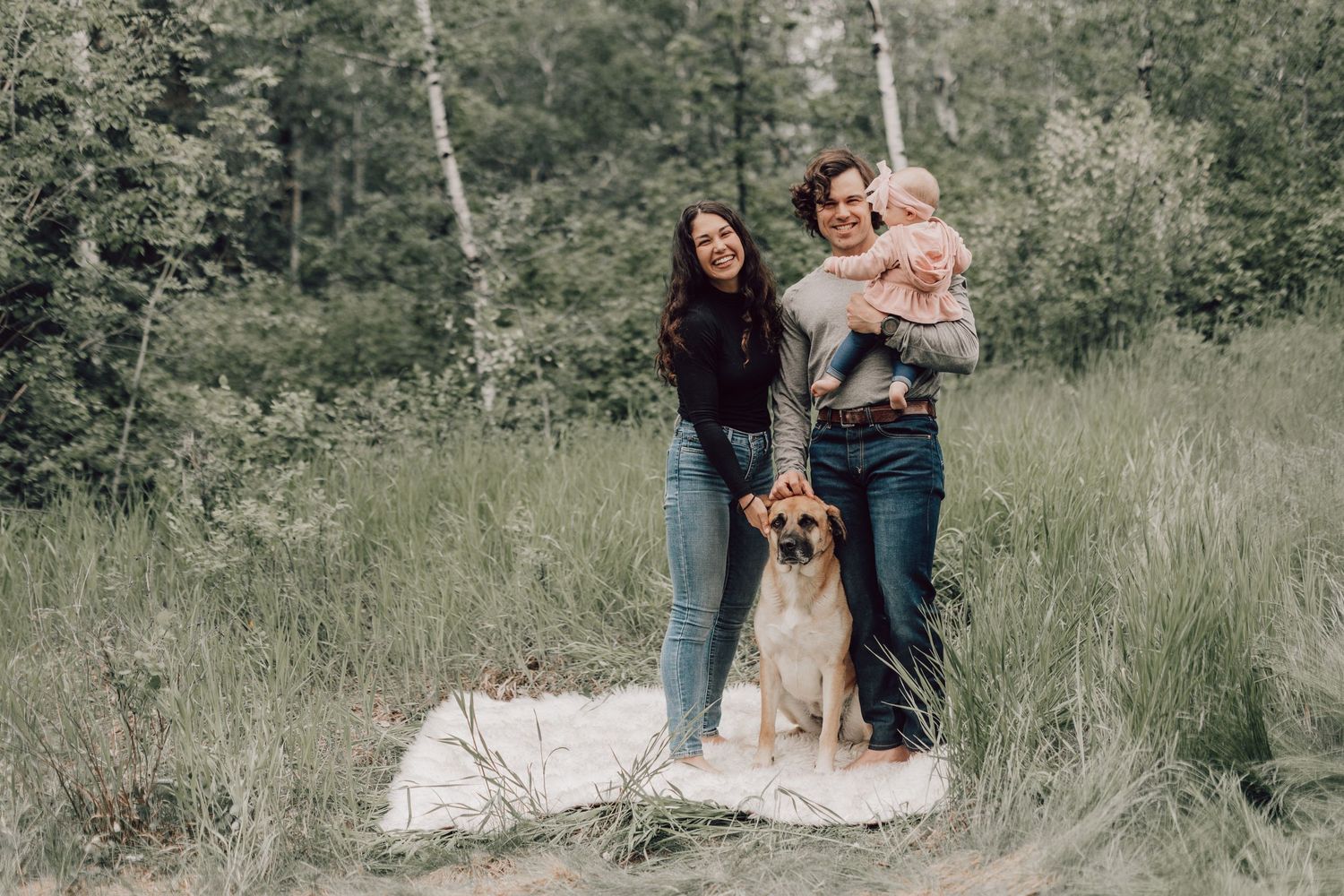 Happy family with dog posing in nature, parents holding baby in green forest meadow.