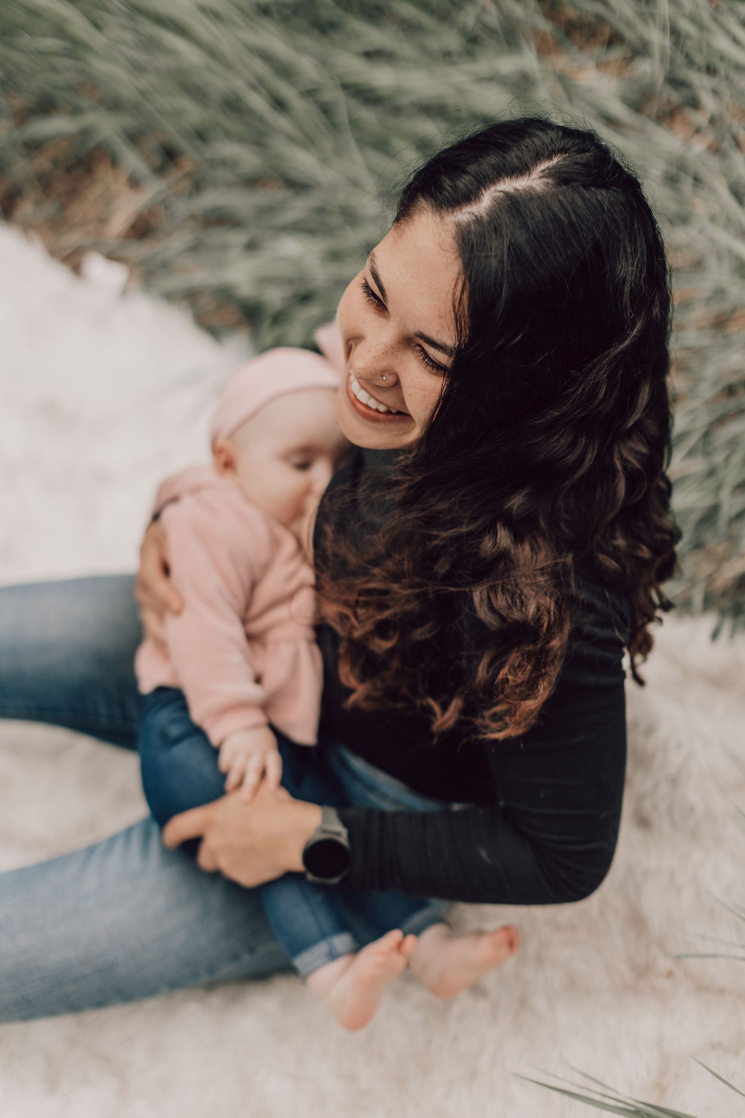 Smiling dark-haired mother sitting outdoors holding infant in pink outfit.