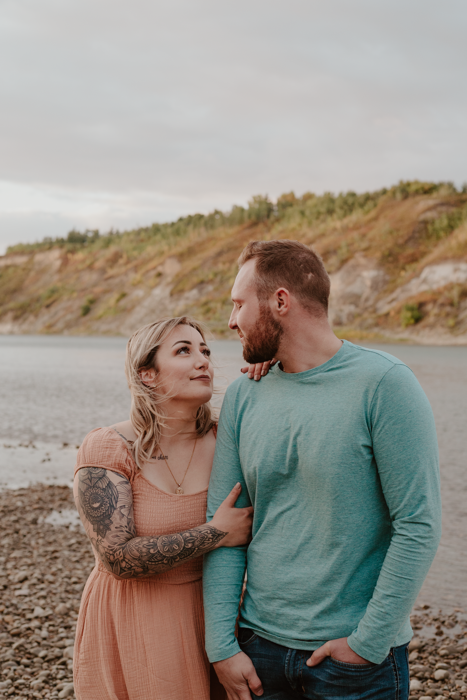 Man and woman standing by a river looking at each other. Man has hands in pocket and she has her hand on his arm.