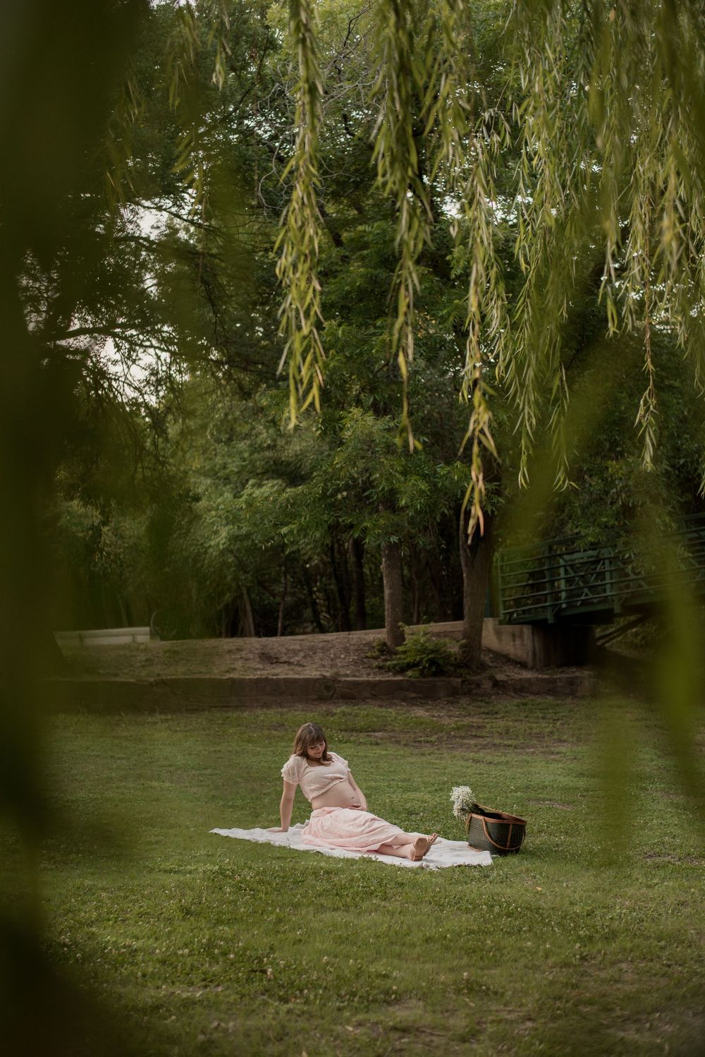 Pregnant woman in pink dress relaxing on white blanket under weeping willow tree in lush green park.