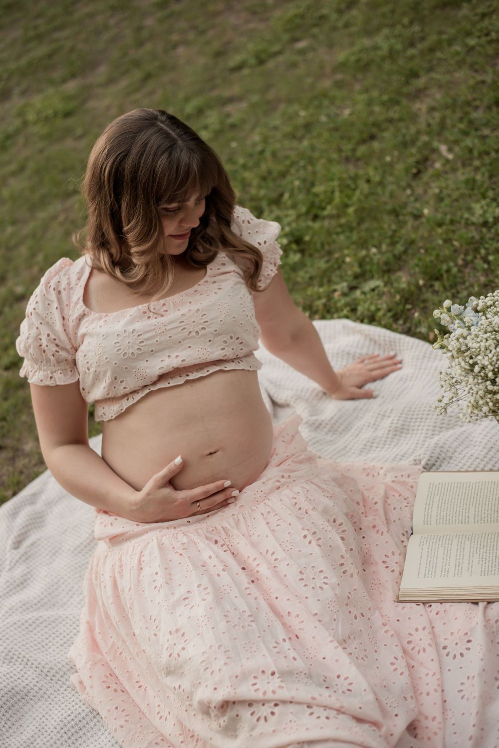 Pregnant woman in pink eyelet crop top and skirt sitting on blanket outdoors, cradling baby bump near open book.