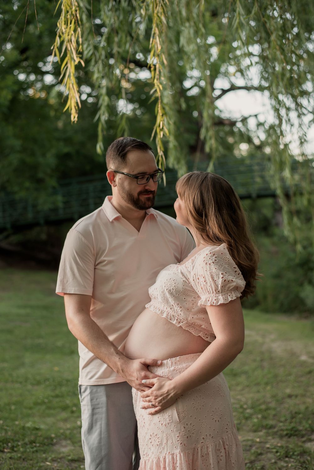 Couple sharing a tender maternity moment outdoors under a willow tree, man cradling pregnant womans belly in a lush green park.