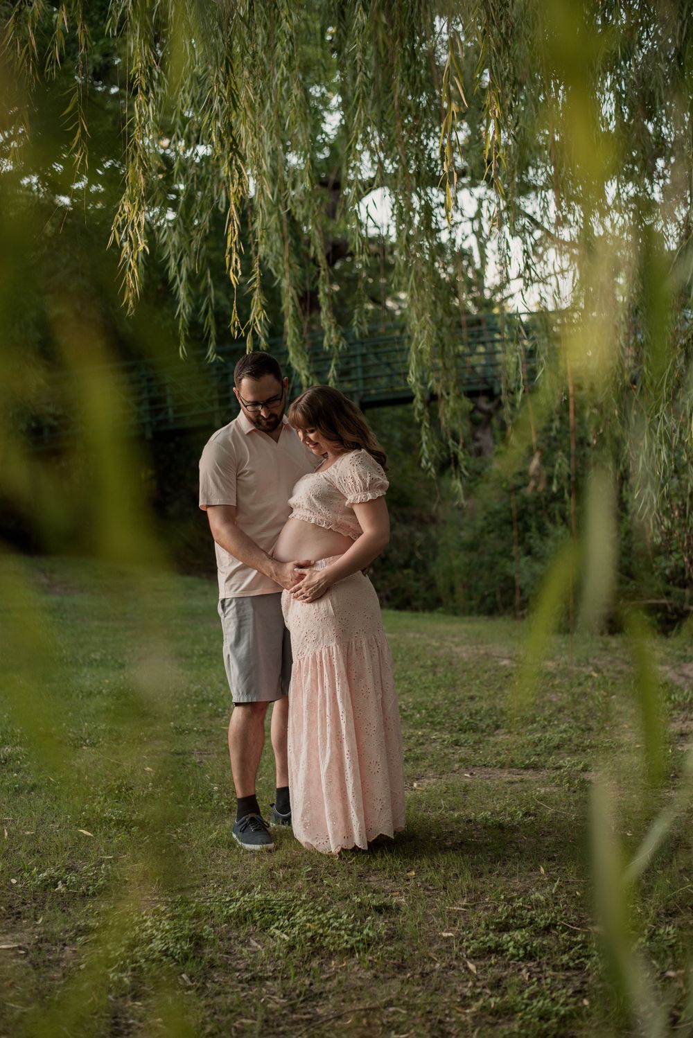 Couple tenderly cradling pregnant belly under weeping willow tree during golden hour maternity photoshoot outdoors.
