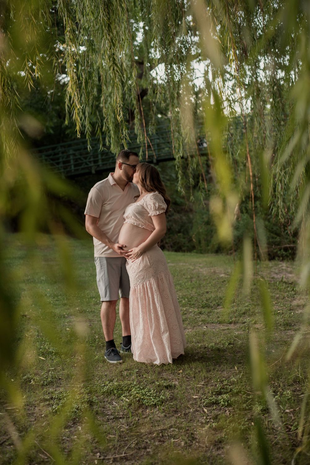 Couple sharing a tender moment under a weeping willow tree during a maternity photo shoot outdoors.