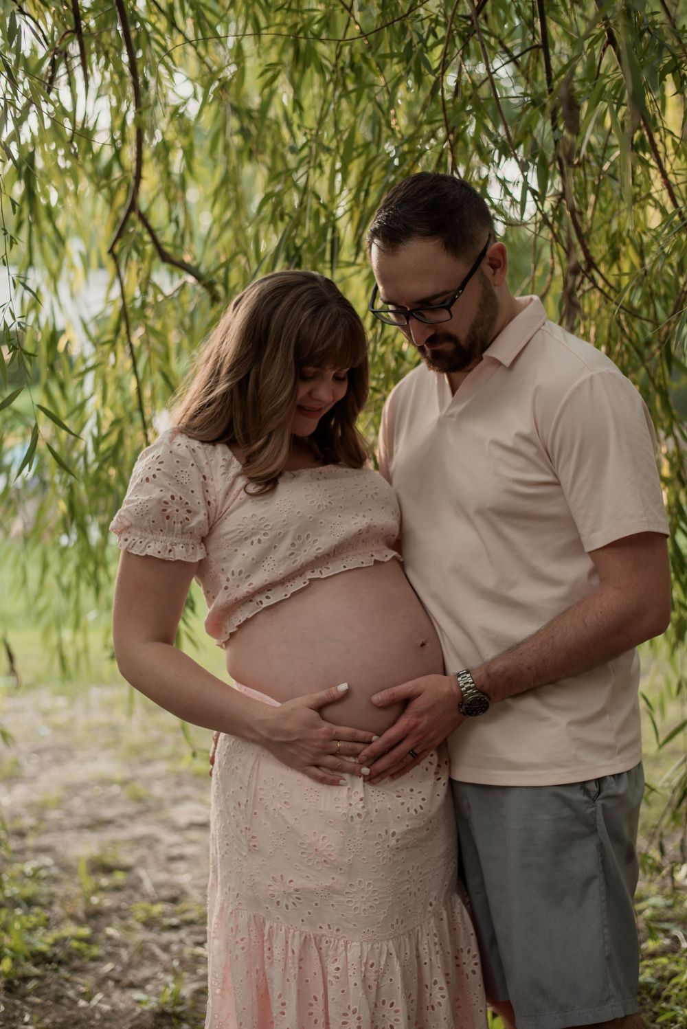 Couple sharing a tender maternity moment under willow trees, cradling pregnant belly outdoors.
