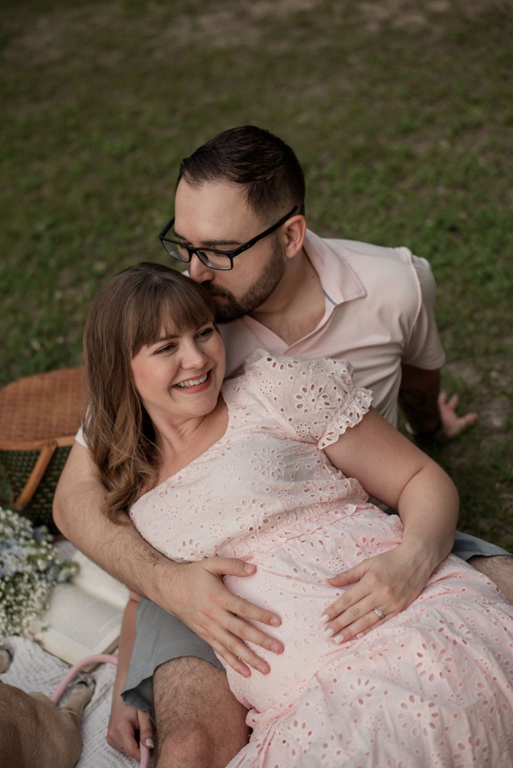 A couple shares a tender moment outdoors, with a man embracing a smiling pregnant woman in a pink lace dress.