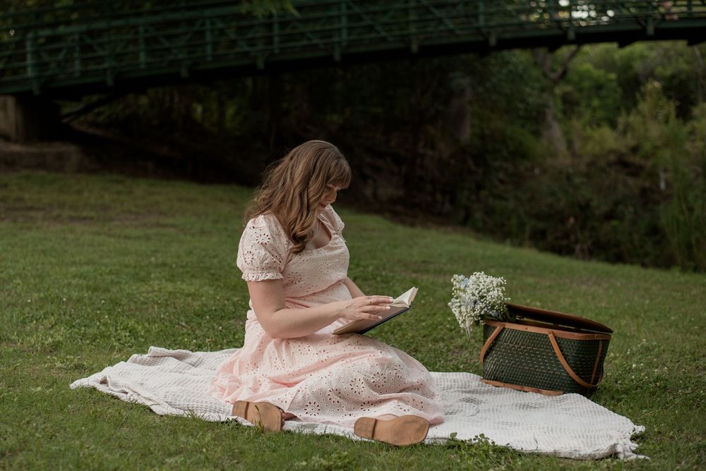 Woman in pink dress sitting on blanket in park, reading a card with picnic basket and flowers nearby.