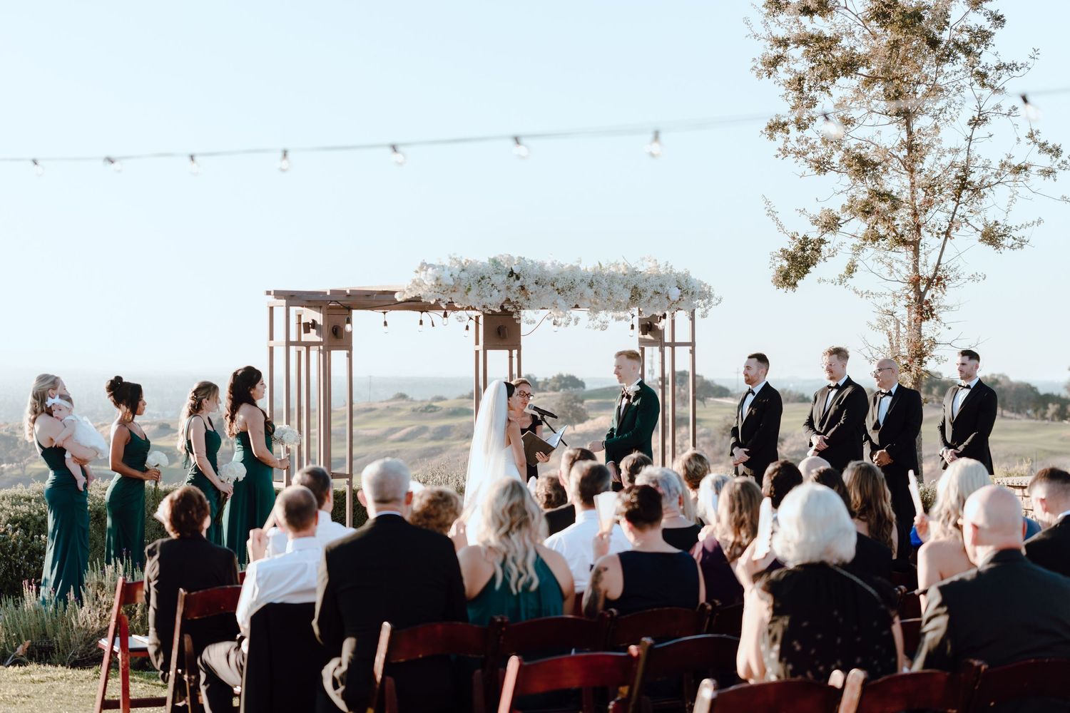 An outdoor wedding ceremony setup with wooden arch and white florals against a scenic backdrop.