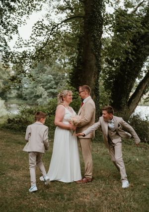 Een vrolijk bruidspaar poseert buiten onder bomen in park Ter Rijst, terwijl kinderen speels naast hen dansen, foto door Lien Photography