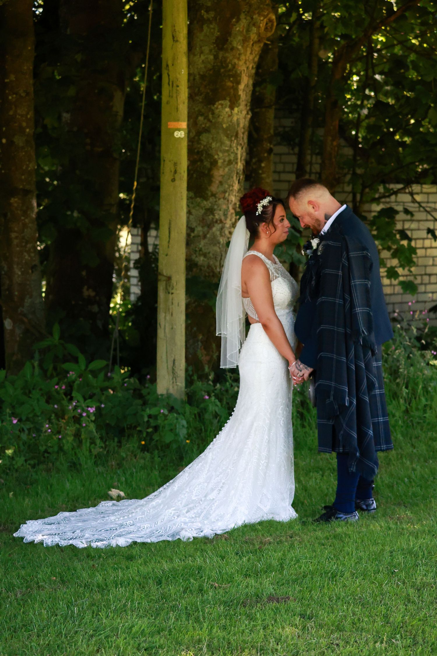 Bride in white lace gown and groom in plaid kilt standing together on grass near trees.