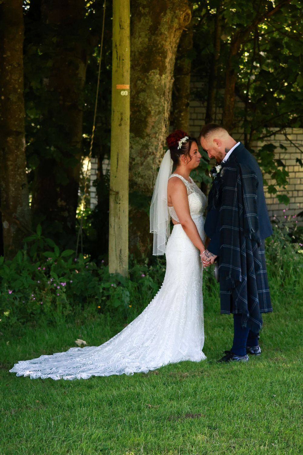 Bride in white lace gown and groom in plaid kilt standing together on grass near trees.