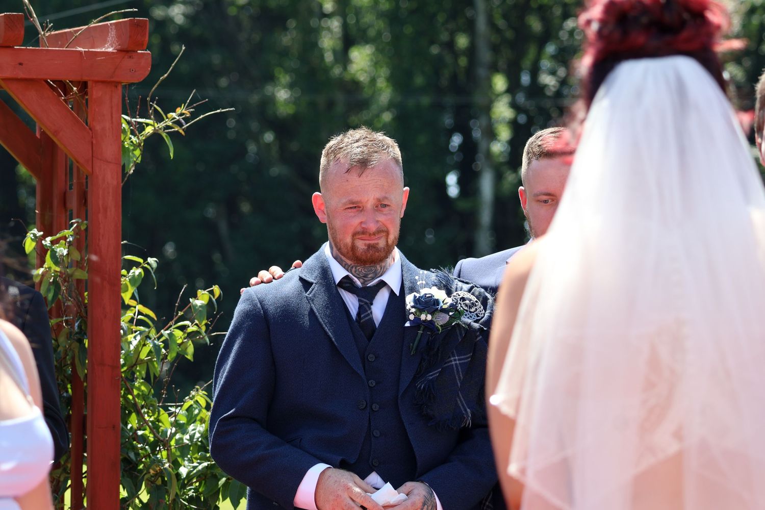 An outdoor wedding ceremony under a wooden pergola surrounded by lush greenery.