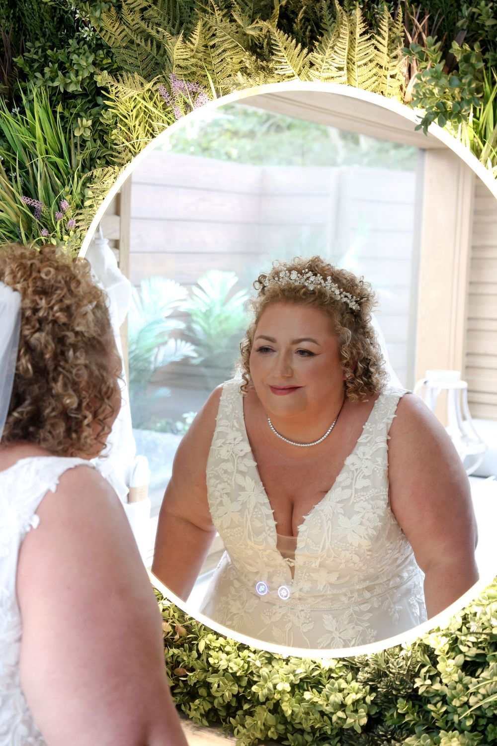 A bride in a white lace dress and pearl necklace looks at her reflection in a circular mirror surrounded by greenery.
