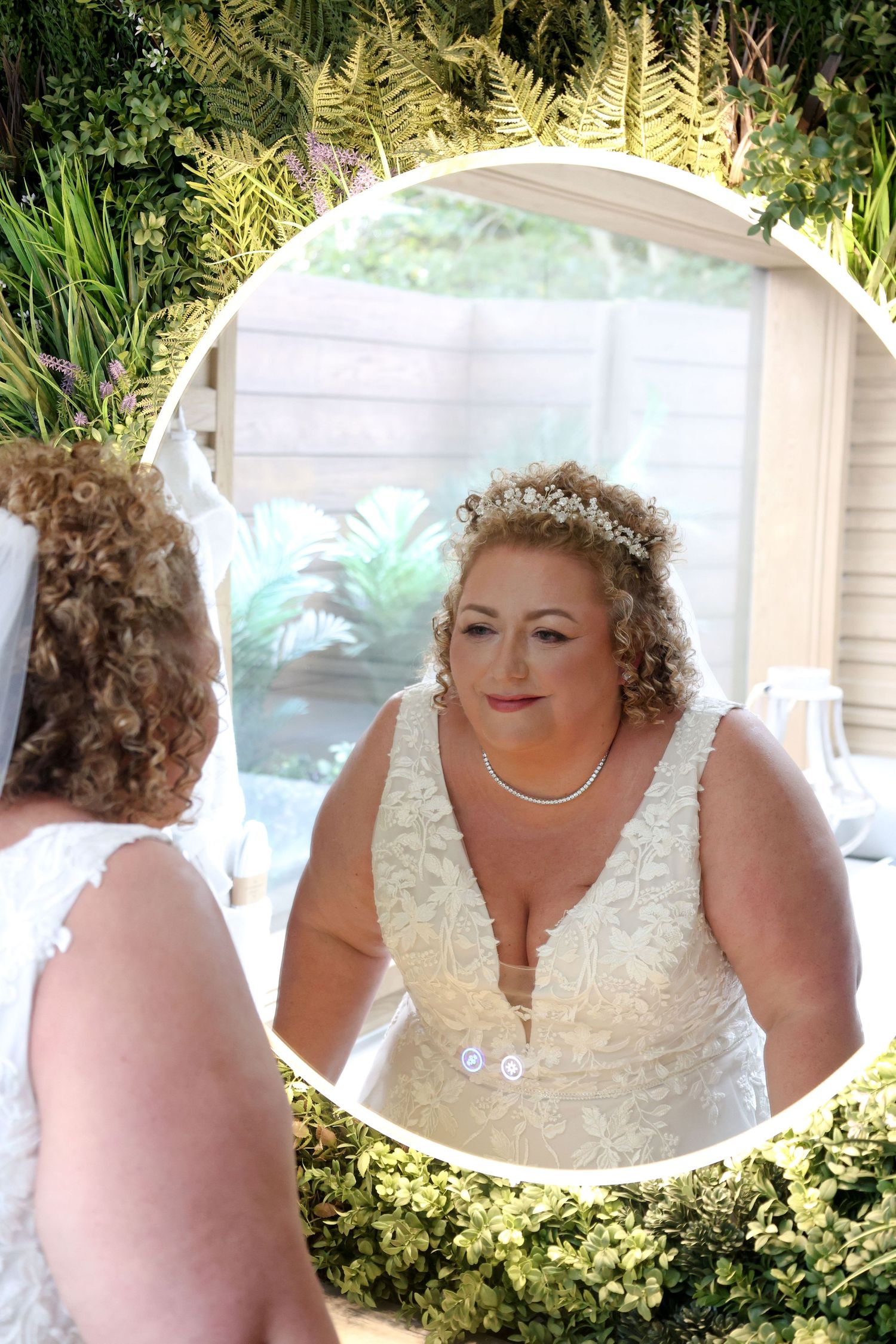 A bride in a white lace dress and pearl necklace looks at her reflection in a circular mirror surrounded by greenery.
