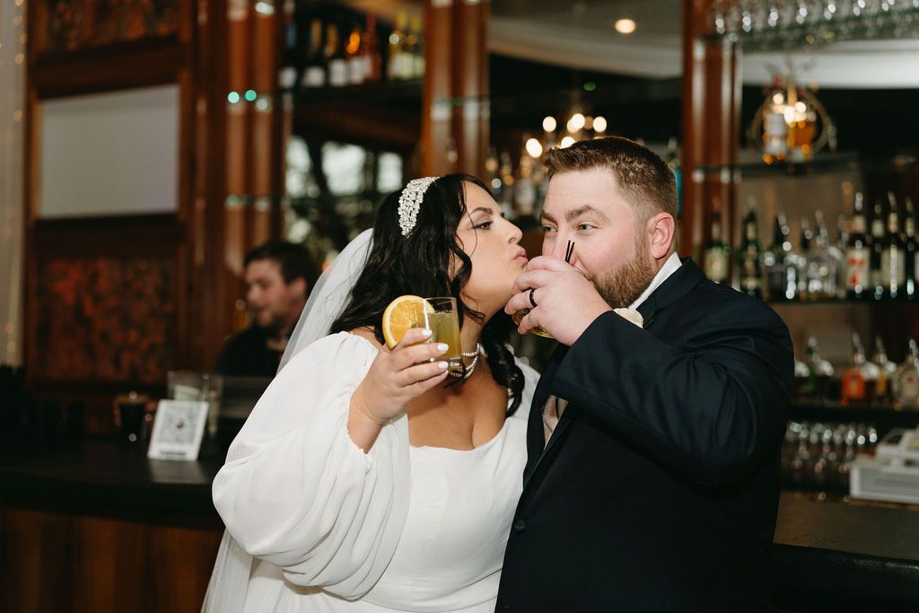 Newlyweds enjoying drinks together at a bar with glass wine racks and warm wooden decor in the background.