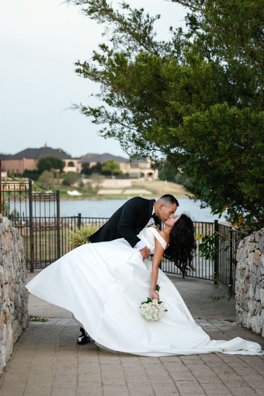 Newlyweds share romantic dip pose on stone pathway with lakeside view in background.
