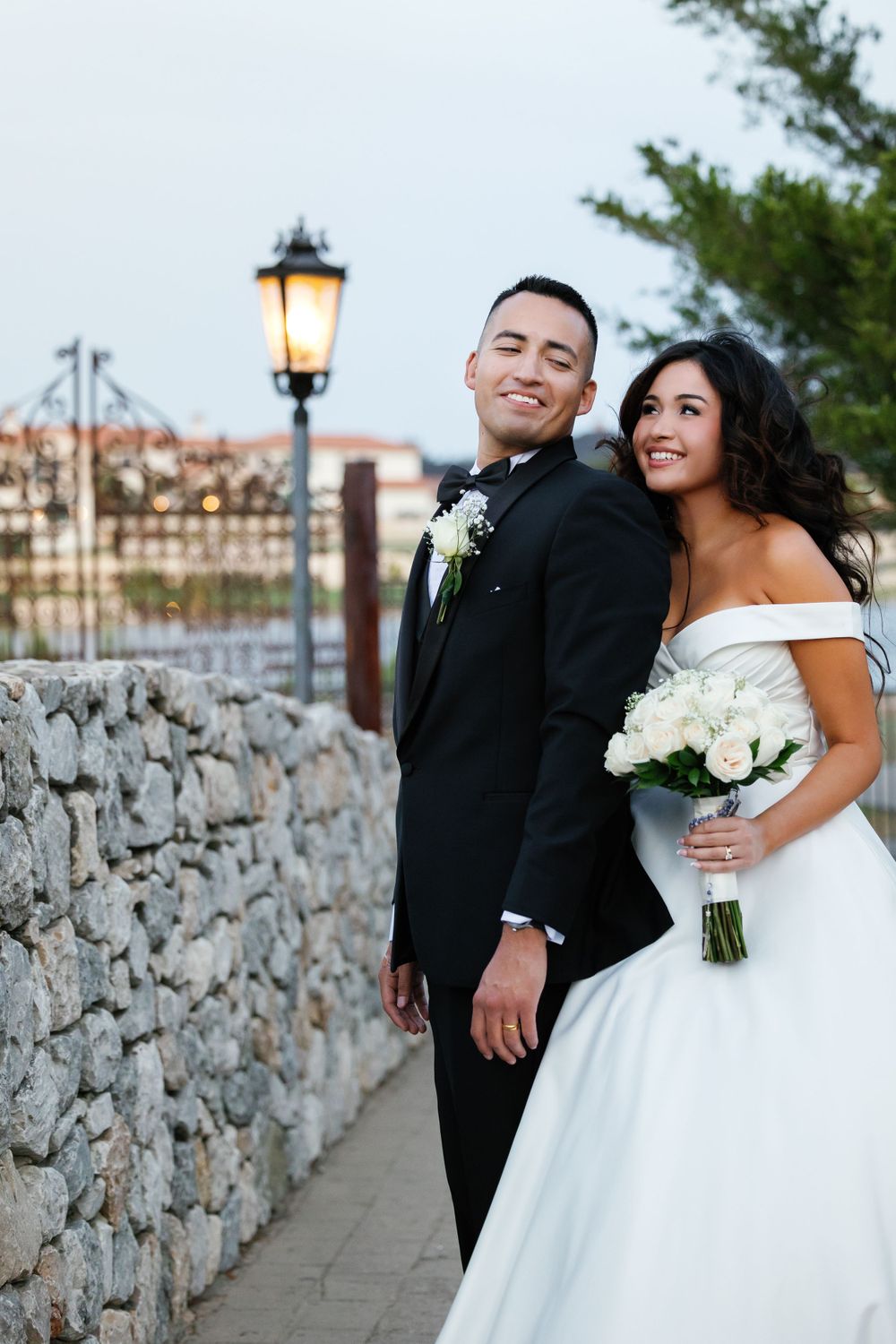 A newlywed couple poses by a stone wall with a glowing lantern at sunset.