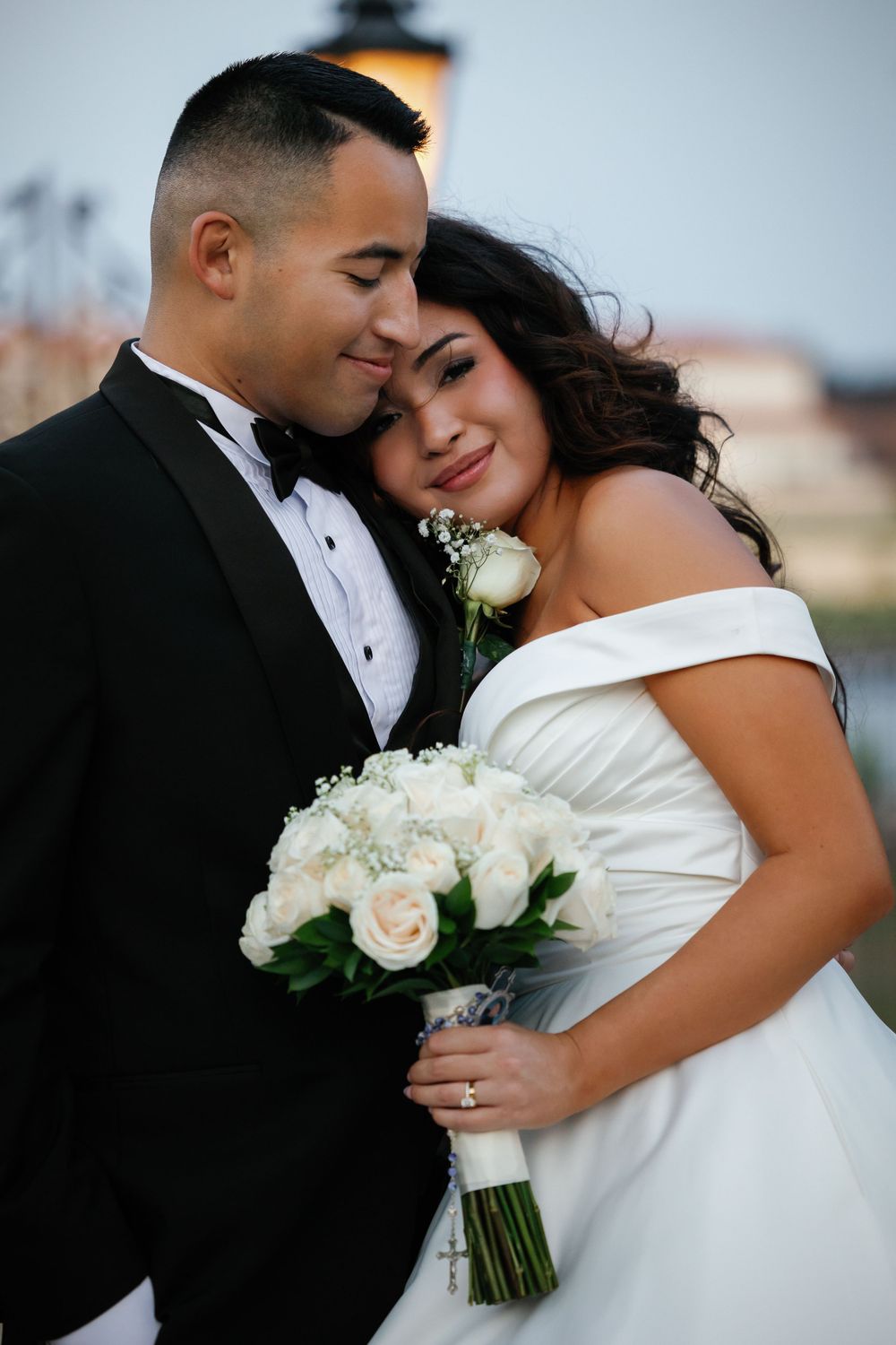 A romantic wedding couple embraces while holding a white rose bouquet during their outdoor portrait session.