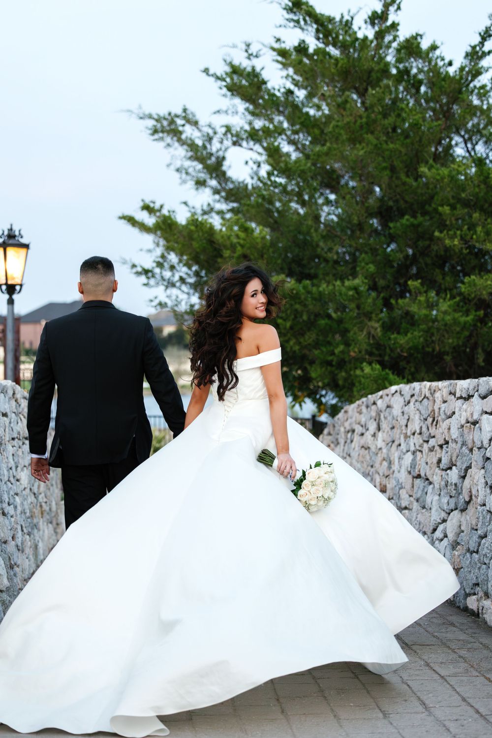Bride's flowing white dress blows dramatically on stone path with water view and trees.