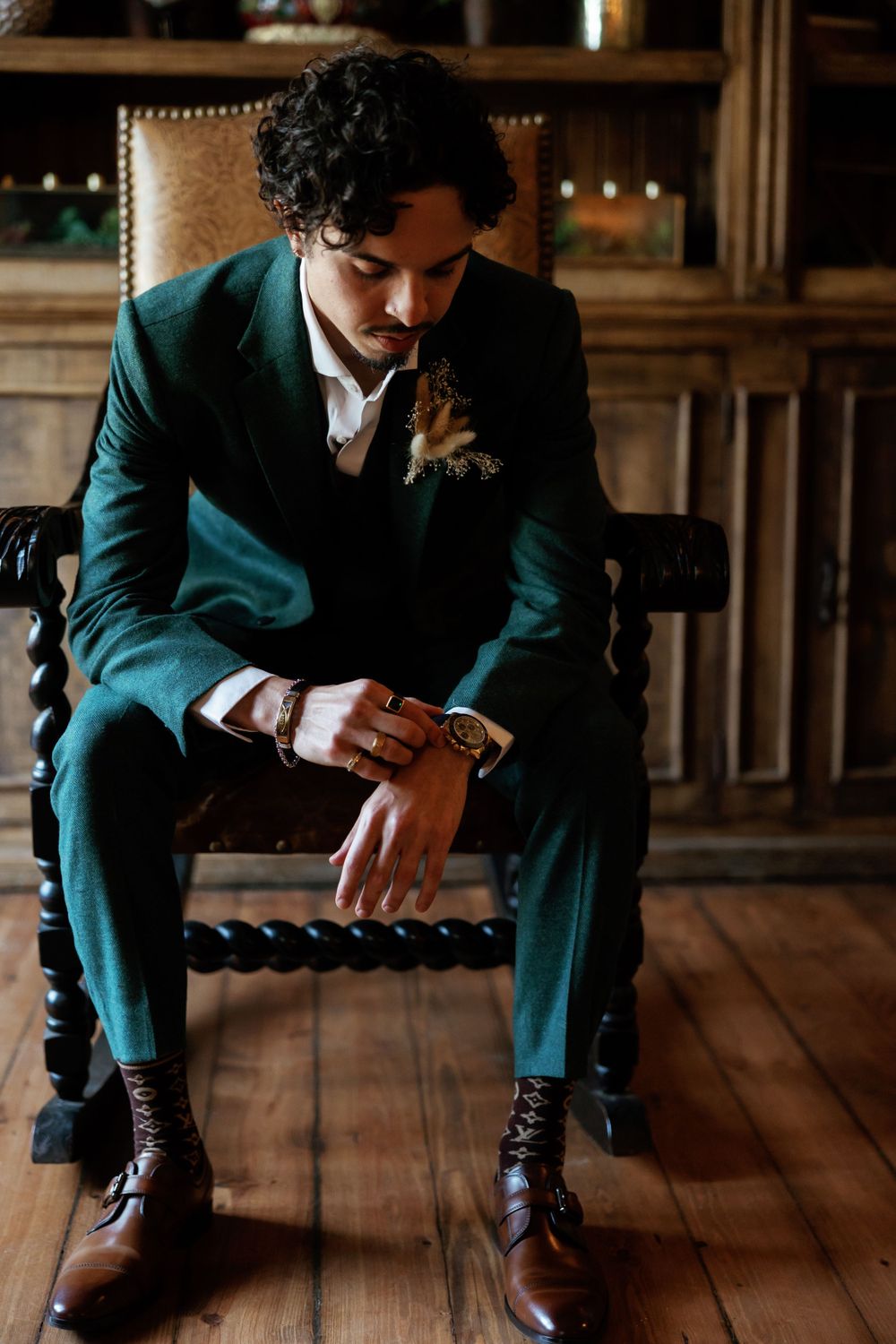 A well-dressed person in an emerald green suit sits contemplatively on a leather chair in a rustic wooden room.