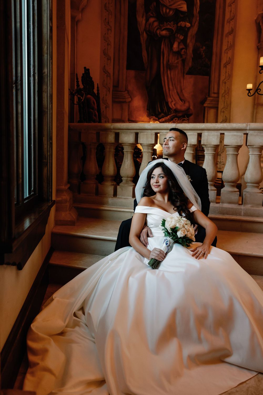 Artistic wedding portrait of couple seated on ornate staircase in warm lighting.