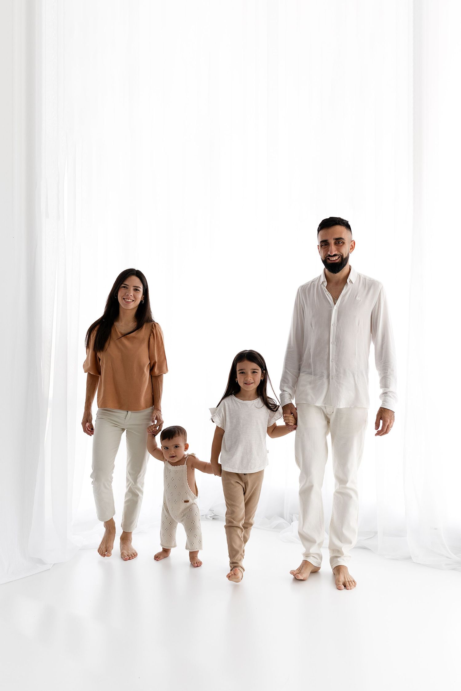 Family wearing coordinated neutral outfits stands together against white backdrop.