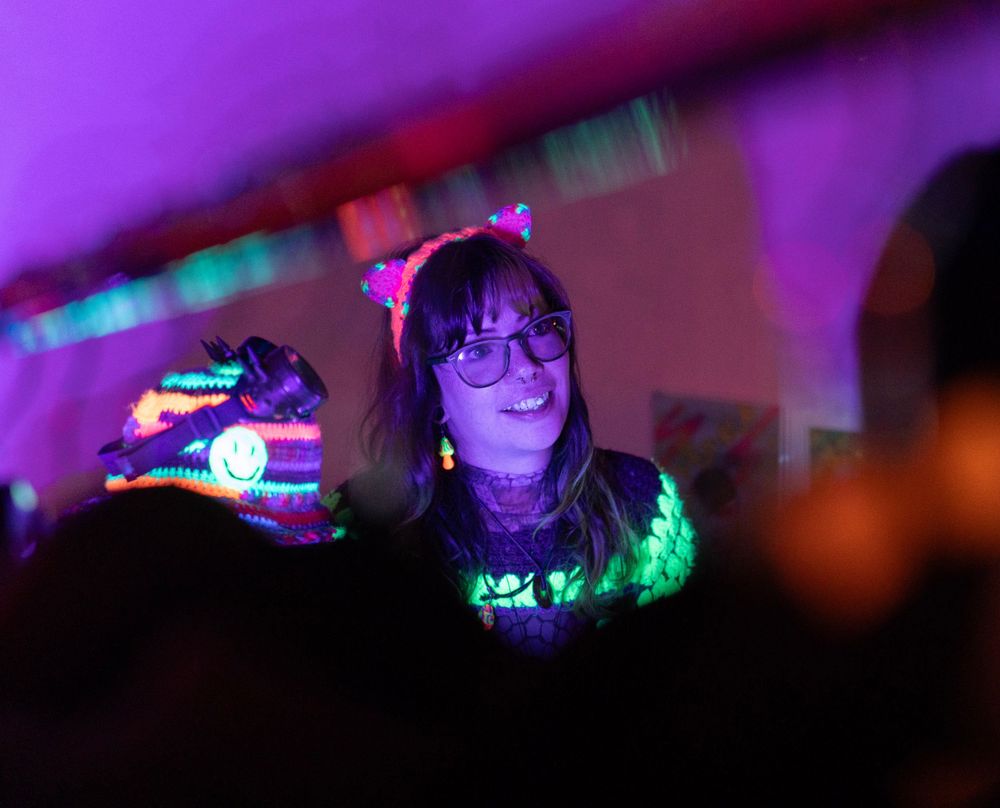woman dressed in crocheted neon costume selling at a handmade art marketplace setup on a table in a dimly neon lit setting. modem