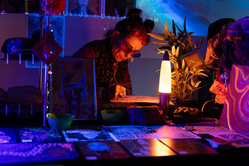 woman Illustrator drawing at a handmade art marketplace setup on a table in a dimly neon lit setting. modem