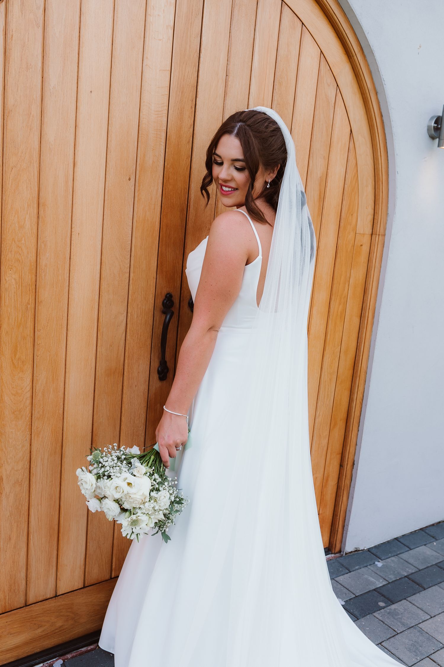 Bride in elegant white dress holds bouquet while standing by wooden arched door. Photographer. Hull. East Yorkshire. Photography