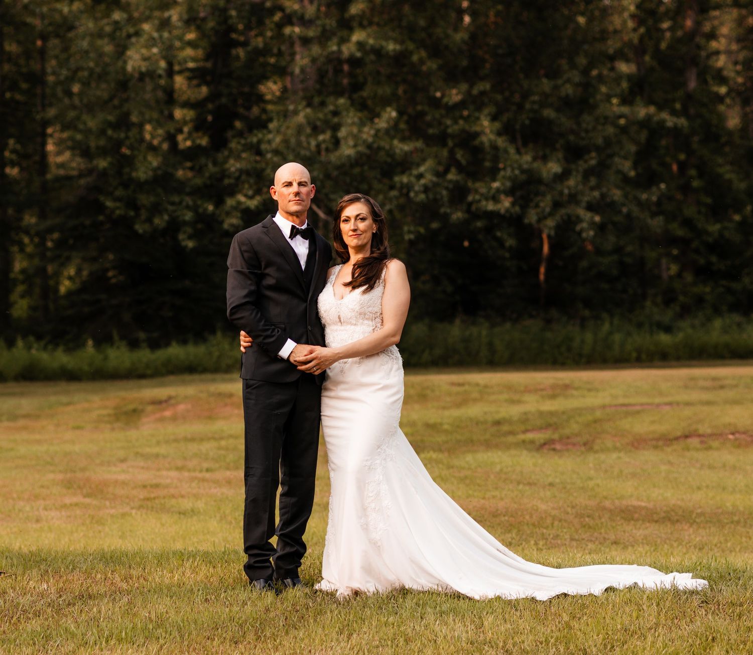 Bride and groom pose together on green grass with tall pine trees in the background.
