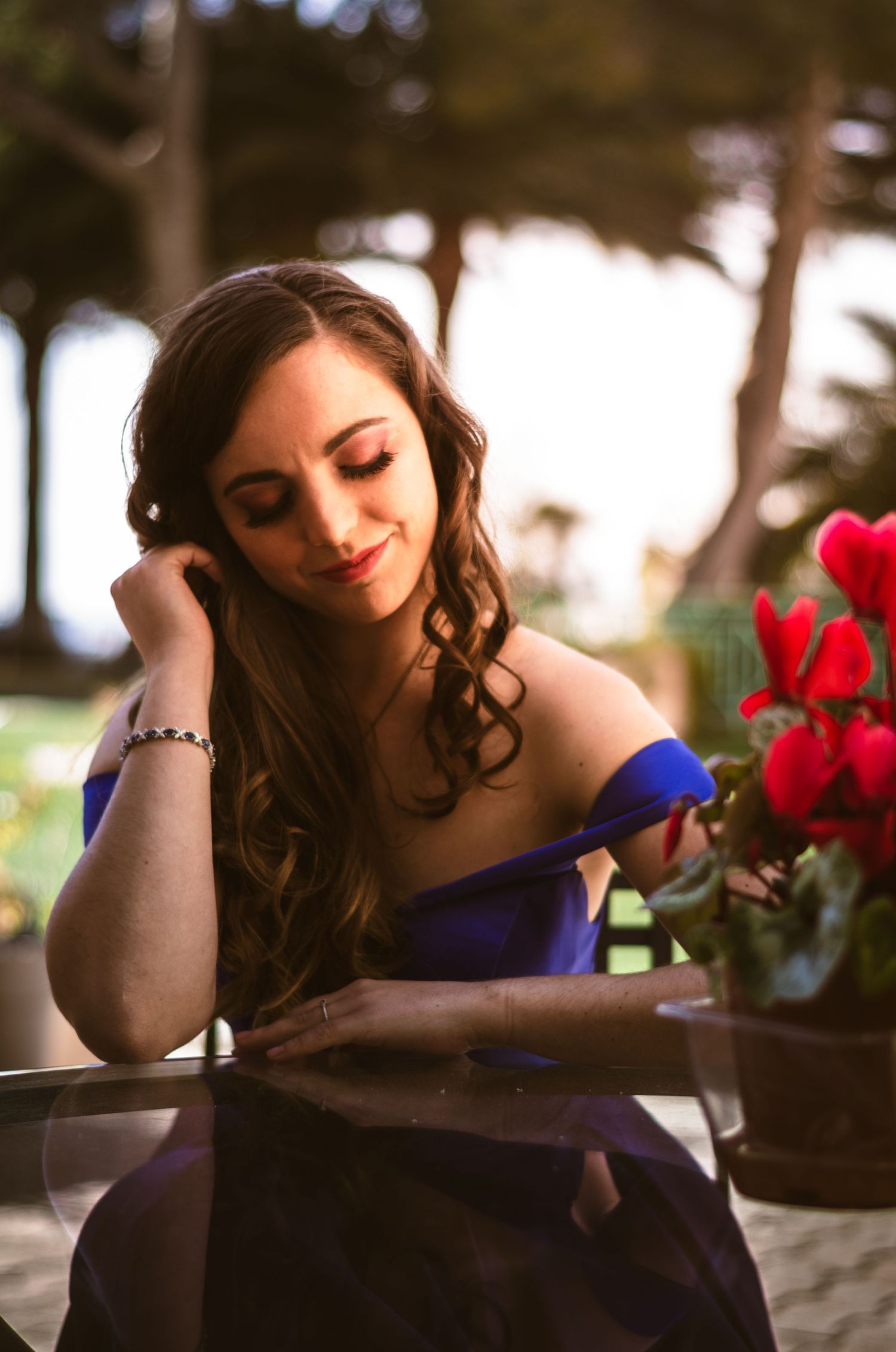 A person in an elegant blue off-shoulder dress sits at a table with red flowers in a romantic outdoor setting.