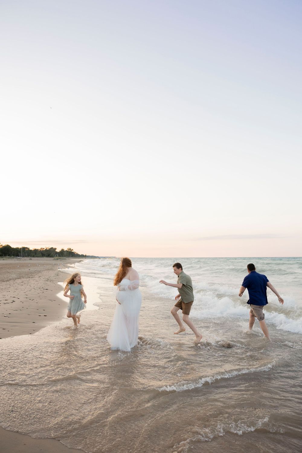 Family of four playing in ocean waves at sunset on a sandy beach, woman in white dress with children and man.