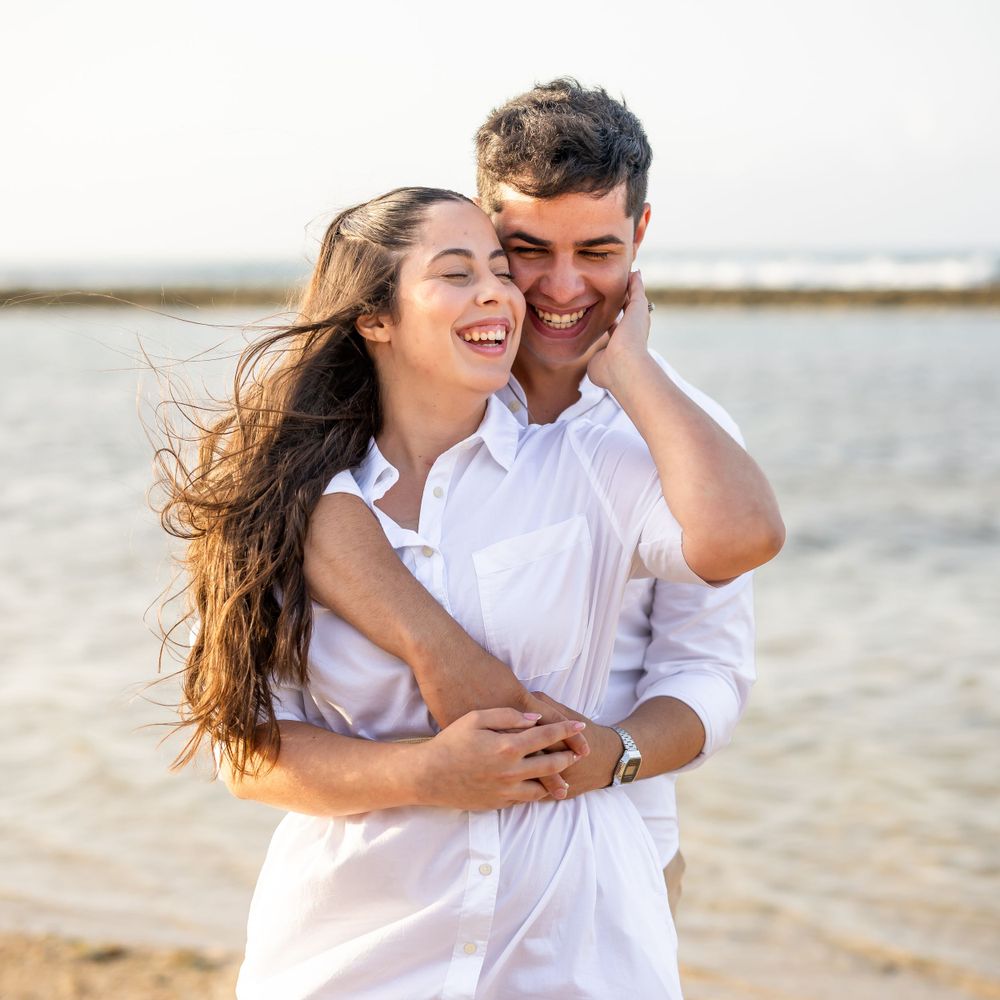 Joyful couple embracing on sunny beach with ocean waves in background.