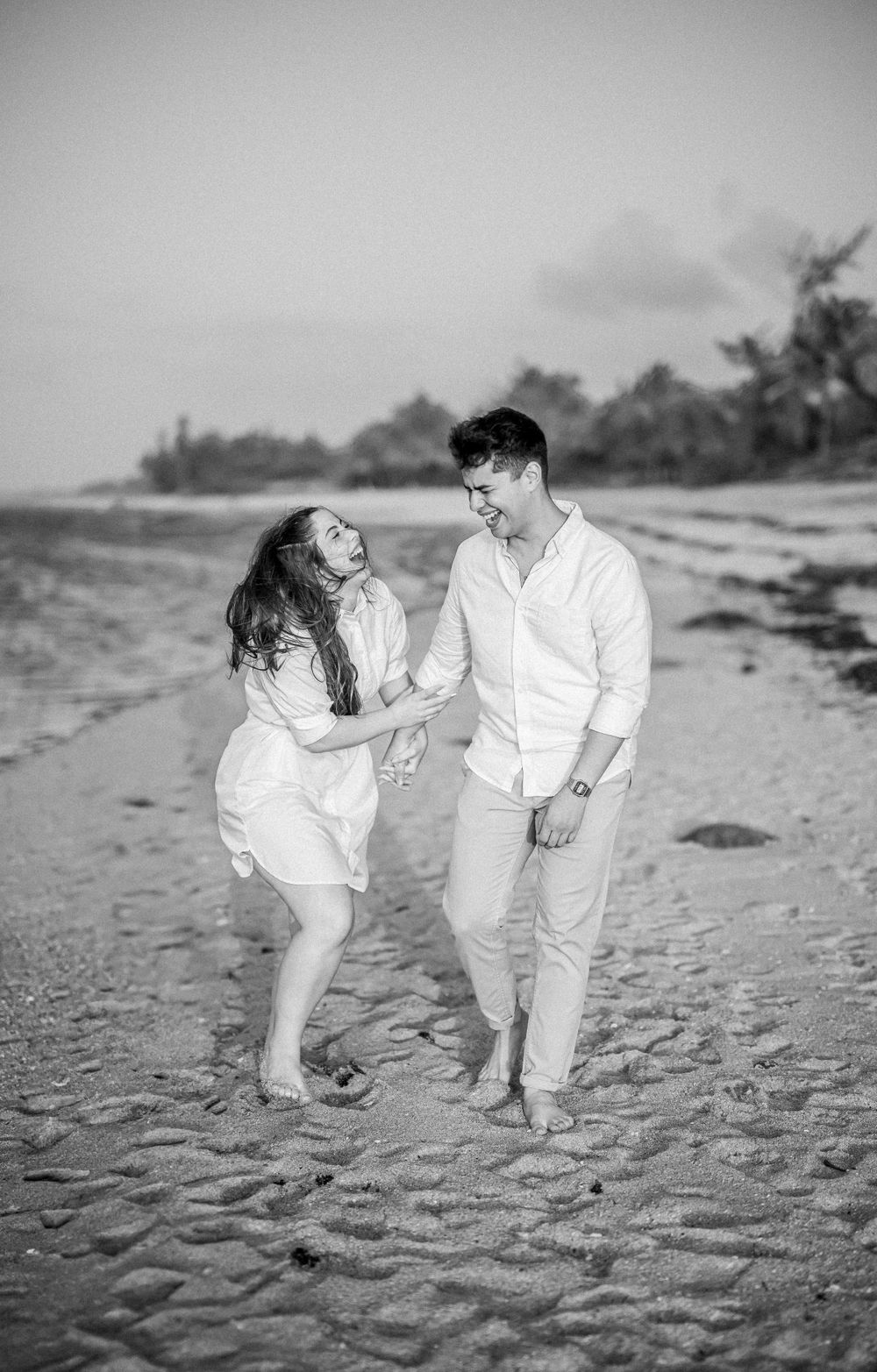 A joyful couple in white outfits laughing together while walking on a tropical beach at sunset.